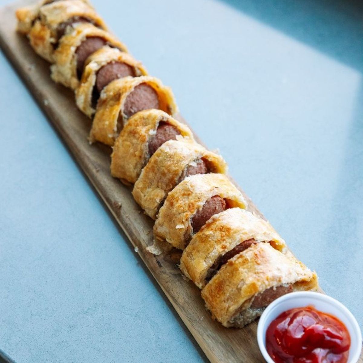 A sliced sausage roll wrapped in golden, flaky pastry is arranged on a wooden board, with a small cup of ketchup at the end for dipping.