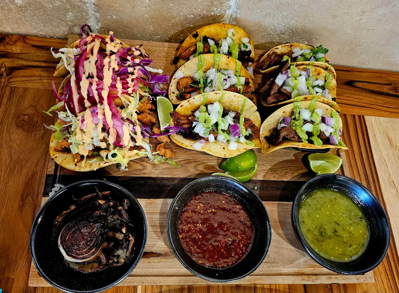 A wooden board displays six tacos with chopped onions and cilantro, three tacos with colorful cabbage and sauce, lime wedges, grilled onions, red salsa, and green salsa in separate bowls.