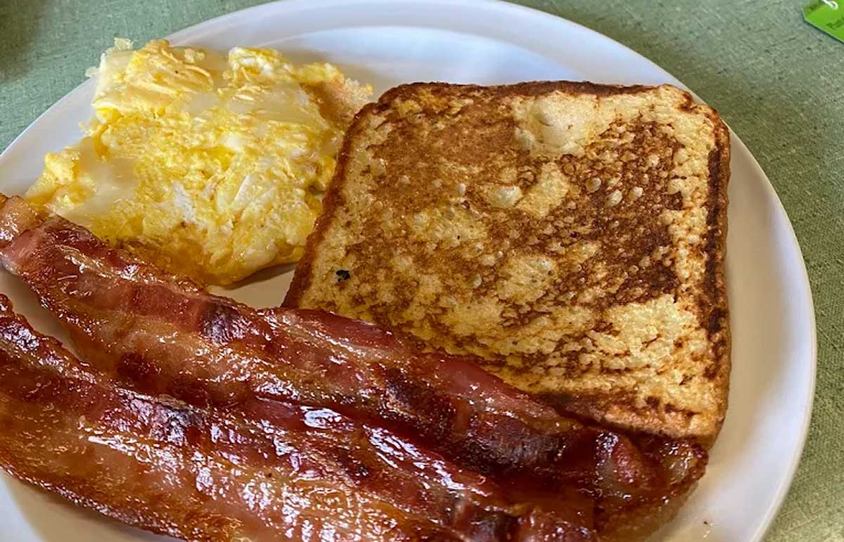 A breakfast plate with two strips of crispy bacon, a piece of French toast, and scrambled eggs—all on a white plate—just like the cheap breakfast in every state.