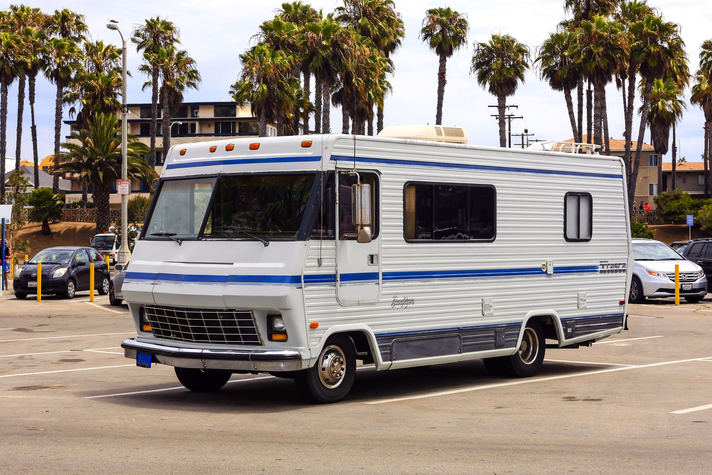 1984 white Itasca Sunflyer RV parked in a parking lot, Santa Monica, California, cars parked in front of large apartment homes and palm trees in the background