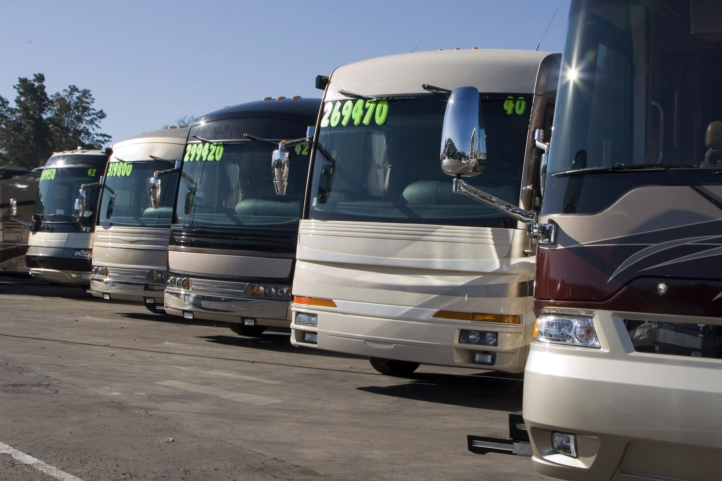 A row of six RVs for sale at an RV dealership, in parking spots, prices on the front window, a blue sky on a sunny day in the background