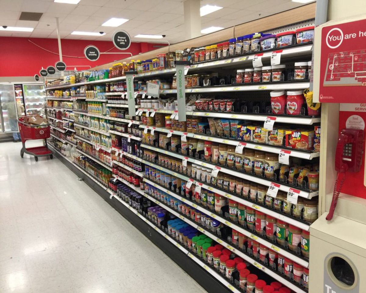 An aisle in the food section at a Target, peanut butter, spreads in the right foreground, salad dressings and other condiments in the background left, with a red shopping cart, freezers line the back, one person getting items from one