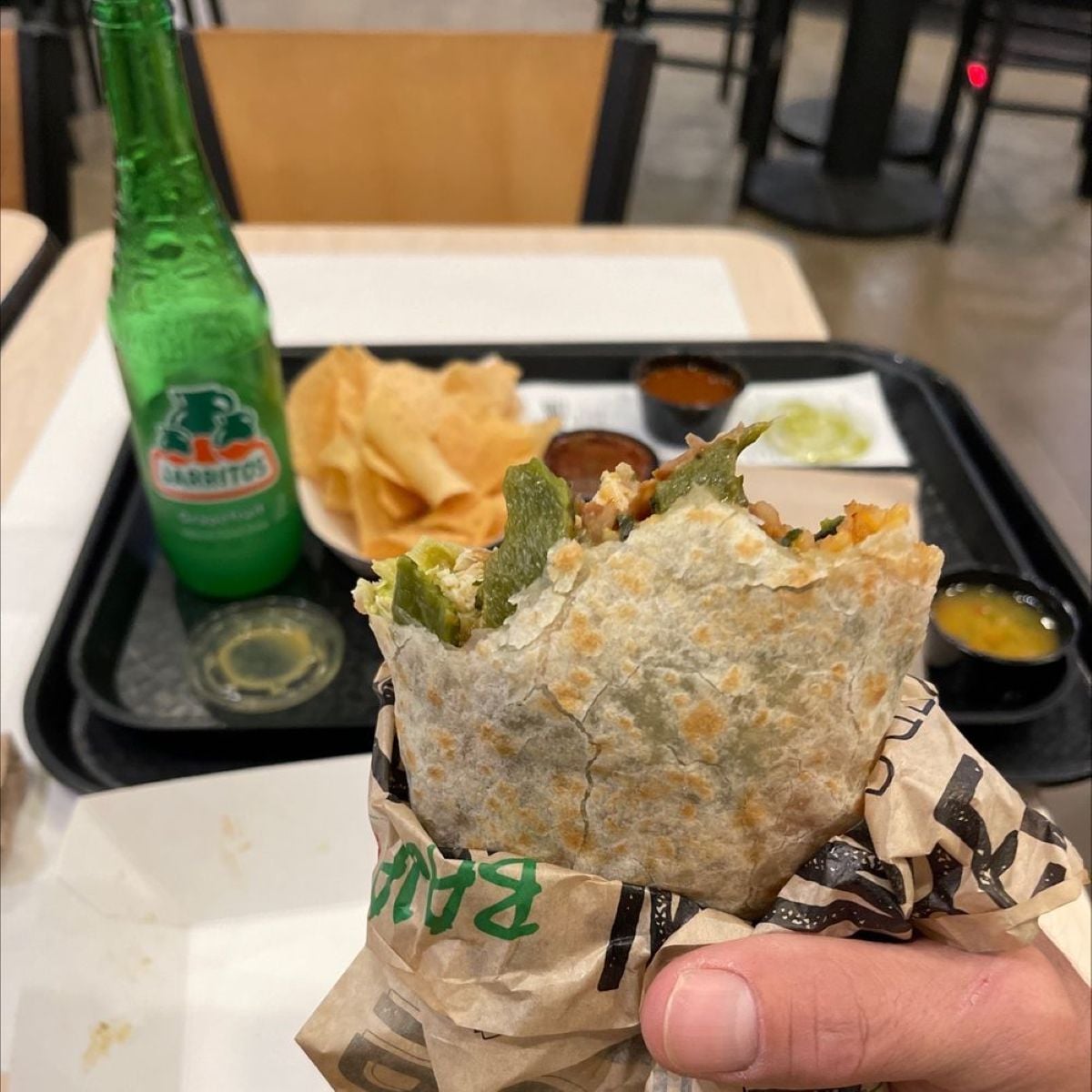 A hand holds a partially eaten burrito wrapped in paper. In the background, there is a green glass bottle, tortilla chips, and trays with various sauces on a table in a casual dining setting.