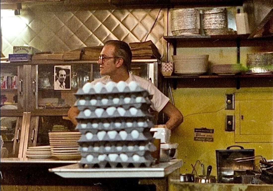 A man with glasses stands in a kitchen behind several trays of eggs, surrounded by shelves with plates and dishes. The background features a photo portrait, a tiled wall, and various kitchen equipment.