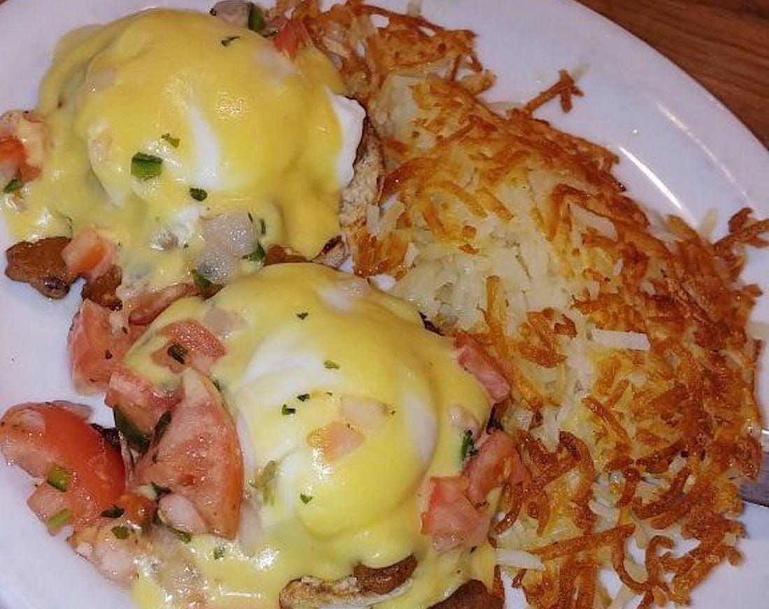 A plate with eggs Benedict topped with hollandaise sauce, fresh diced tomatoes, and herbs, served alongside a portion of crispy golden hash browns.