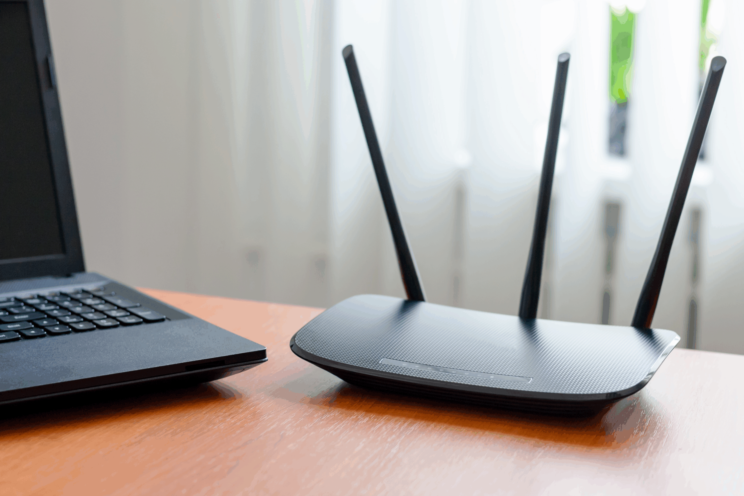 Focus on a black wireless wi-fi router on a wooden desk, a laptop on the left with a semi-transparent white curtains, bright sunlight coming through window