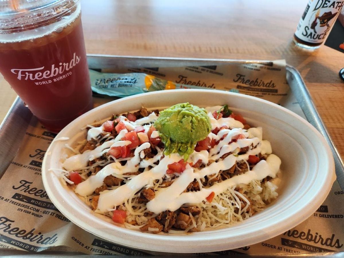 A bowl from Freebirds filled with rice, shredded meat, cheese, diced tomatoes, guacamole, and drizzled with white sauce, next to a red drink cup and a bottle of "Death Sauce" hot sauce on a tray.