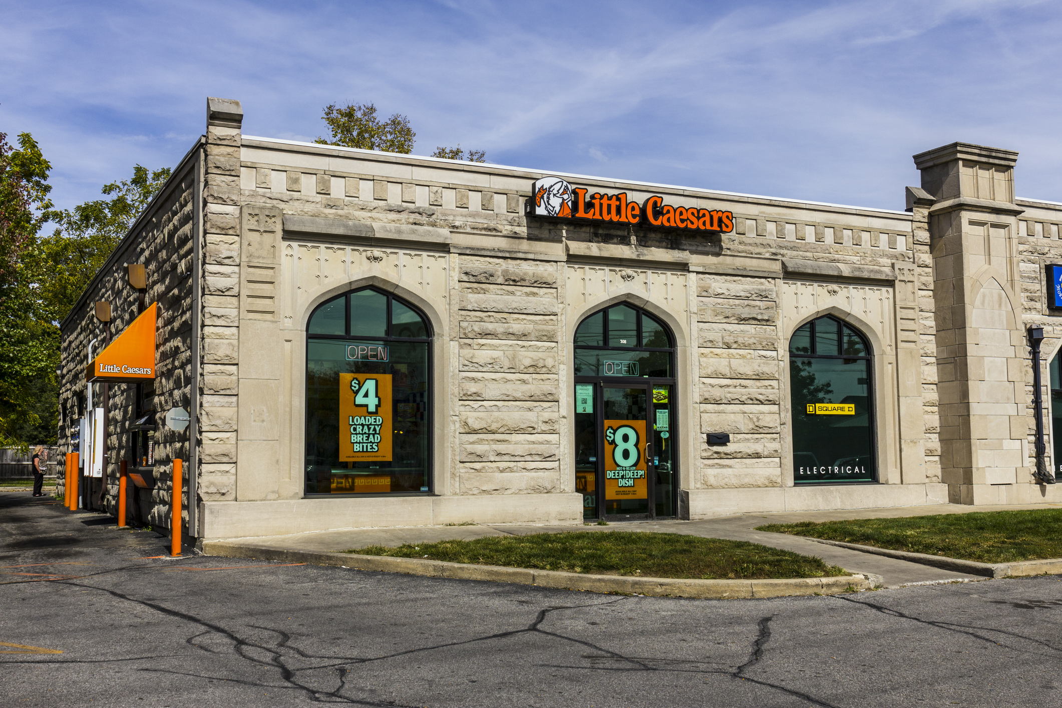 Front exterior of Little Caesars Pizza Restaurant, Kokomo, Indiana, drive-thru on the left, castle-like building, surrounded by road, with trees in the background, against a blue sky