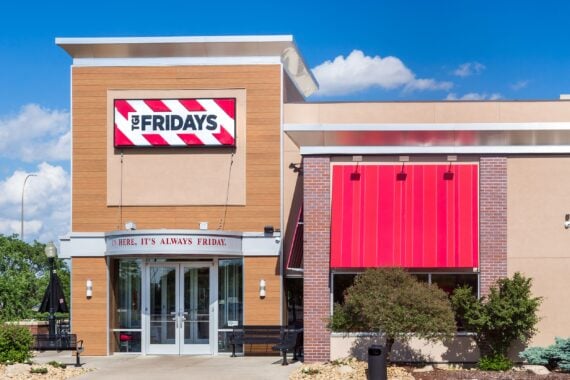 TGI Fridays restaurant exterior with red striped sign, glass entrance doors, bold red awnings, and landscaped bushes under a blue sky with clouds.