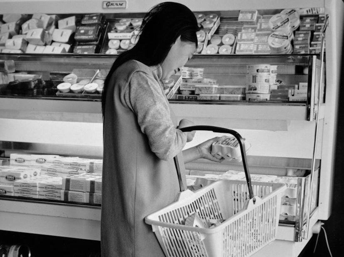 A woman with long dark hair holds a shopping basket and examines a product in the dairy section of a grocery store, surrounded by various packaged dairy items in a refrigerated display.