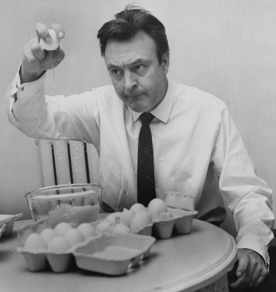 A man in a dress shirt and tie cracks an egg into a bowl at a table covered with several cartons of eggs, looking focused and serious.