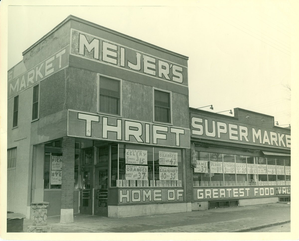 Black and white photo of Meijer’s Thrift Super Market, with large painted signs on the building and produce specials displayed in the windows, advertising items like celery and oranges.