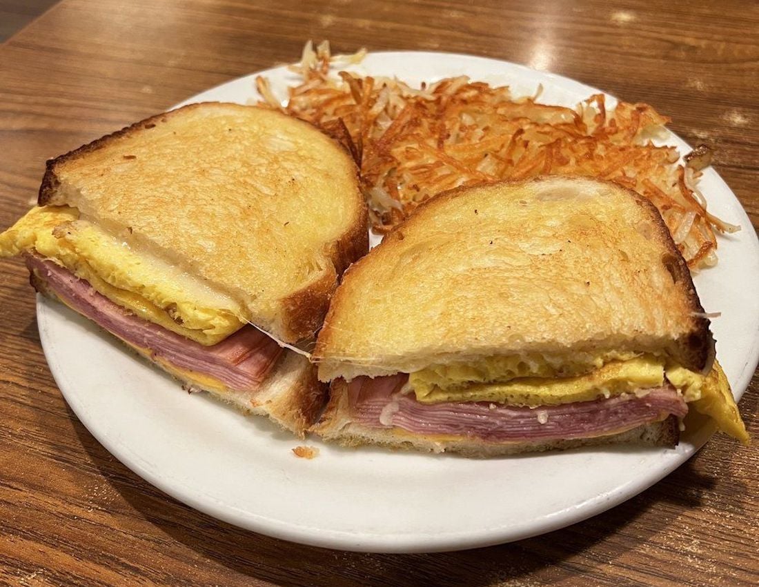 A Denny's plate with a toasted sandwich filled with ham and egg, cut in half, served alongside a portion of crispy hash browns on a wooden table.