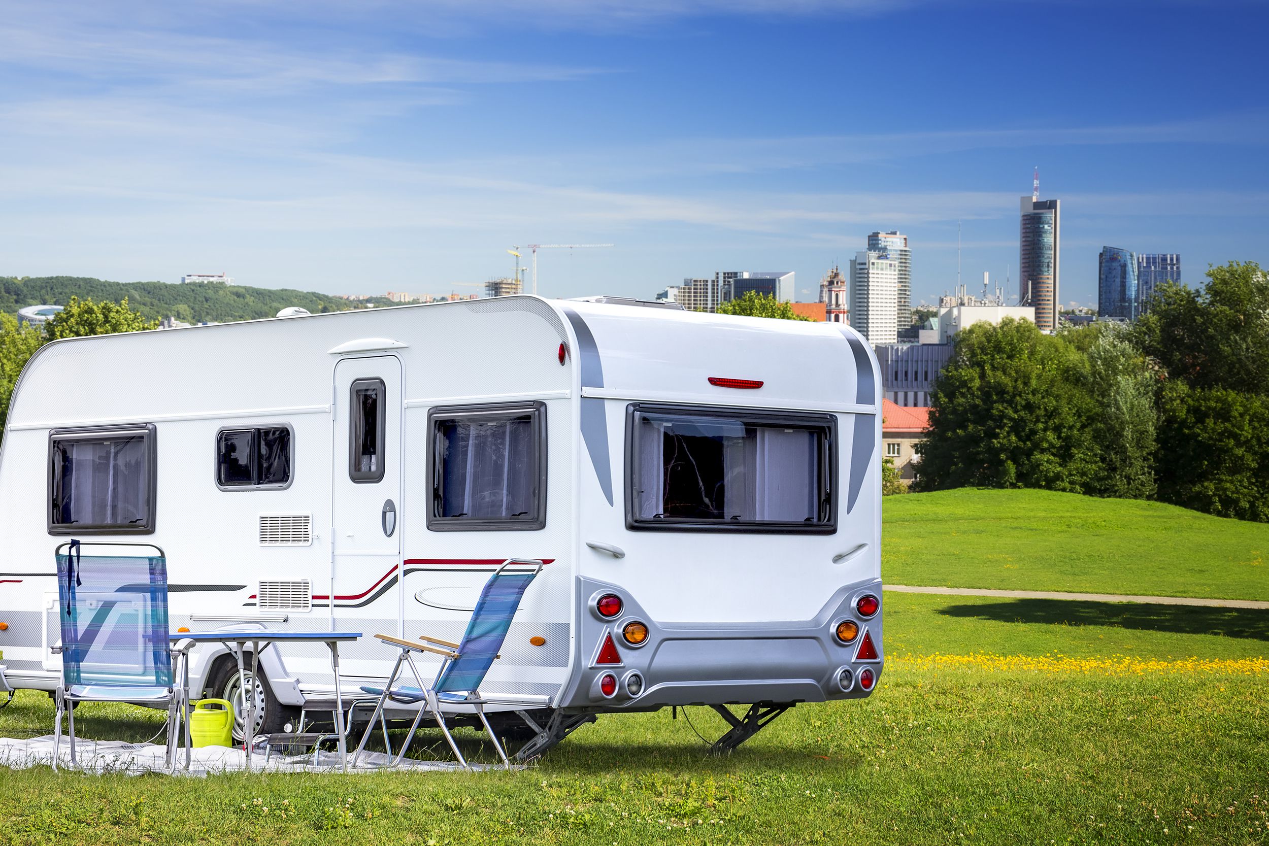 RV parked on grass of a park, on the left foreground, skyscrapers of the downtown city in the background, a blue sky with white clouds, on a sunny day