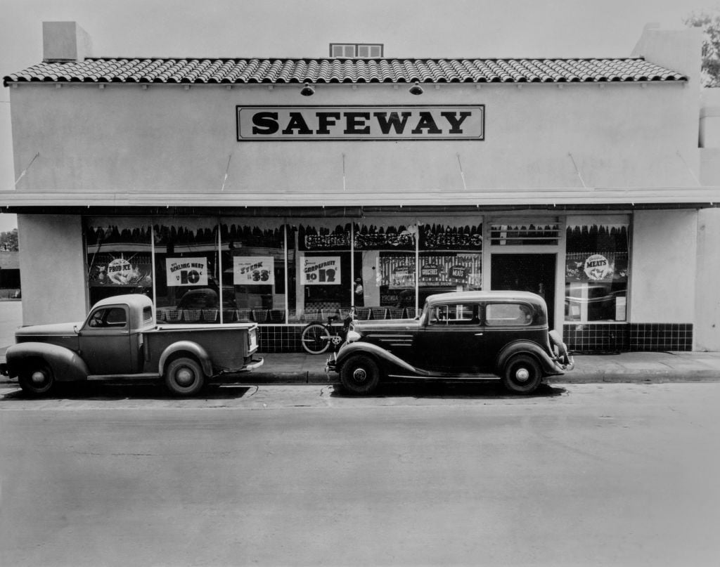 Black and white photo of a vintage Safeway grocery store with two classic cars parked in front and large sale signs displayed in the windows.
