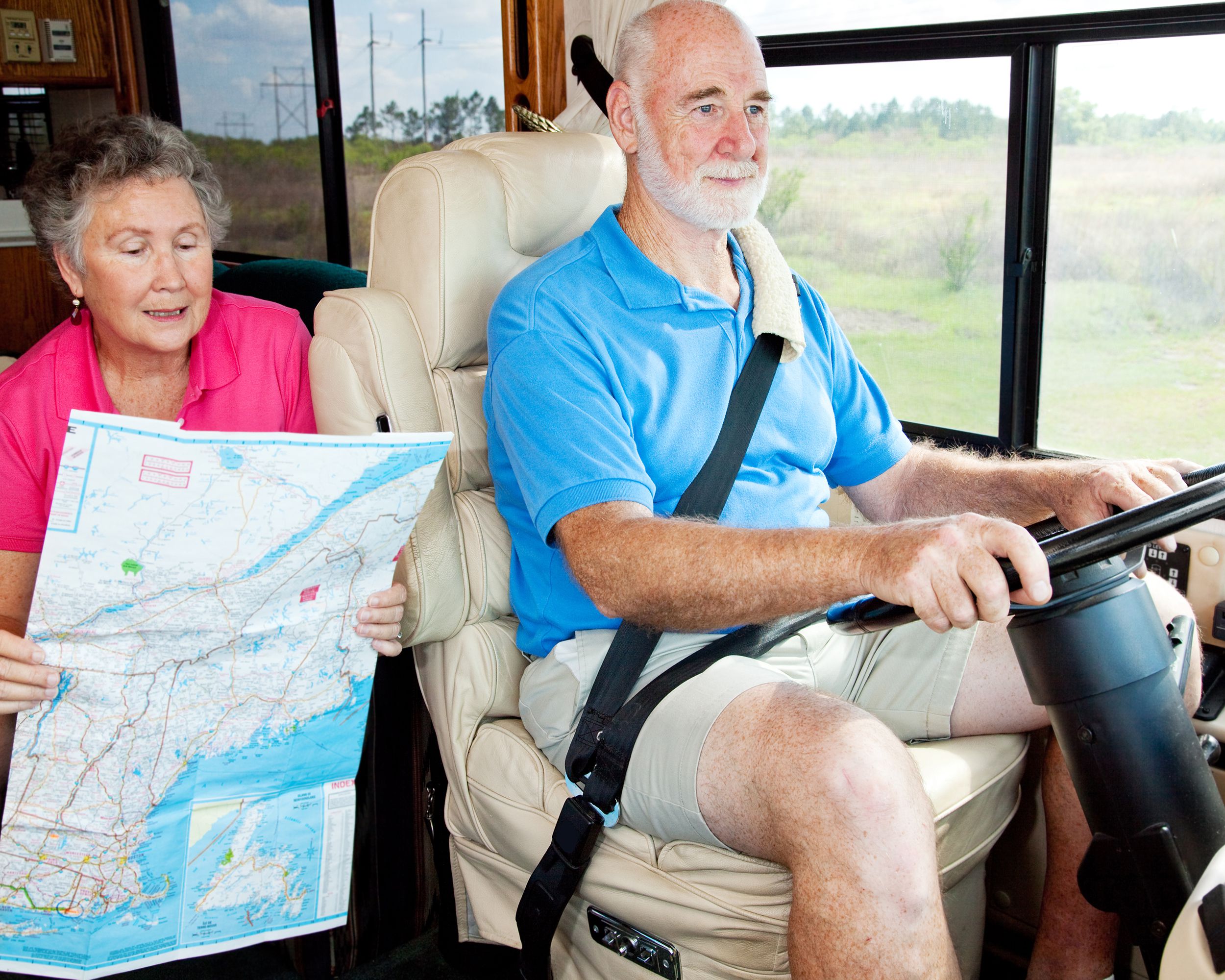 Senior couple in an RV, male is driving on the right and female is giving directions while looking at a map on the left, grassy meadows outside the window