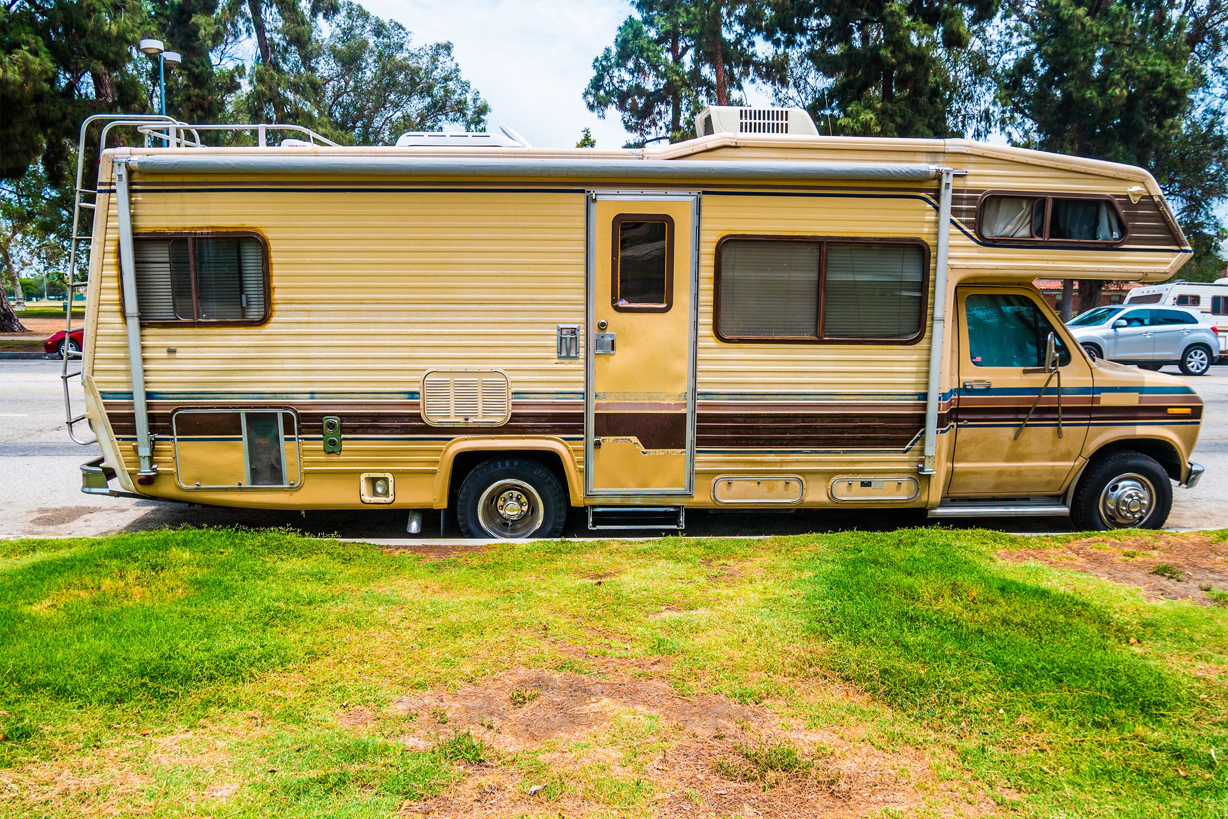 Side-view of beige with dark brown lines RV parked in a parking lot in North Hollywood, lawn in the foreground, cars in the parking lot and trees