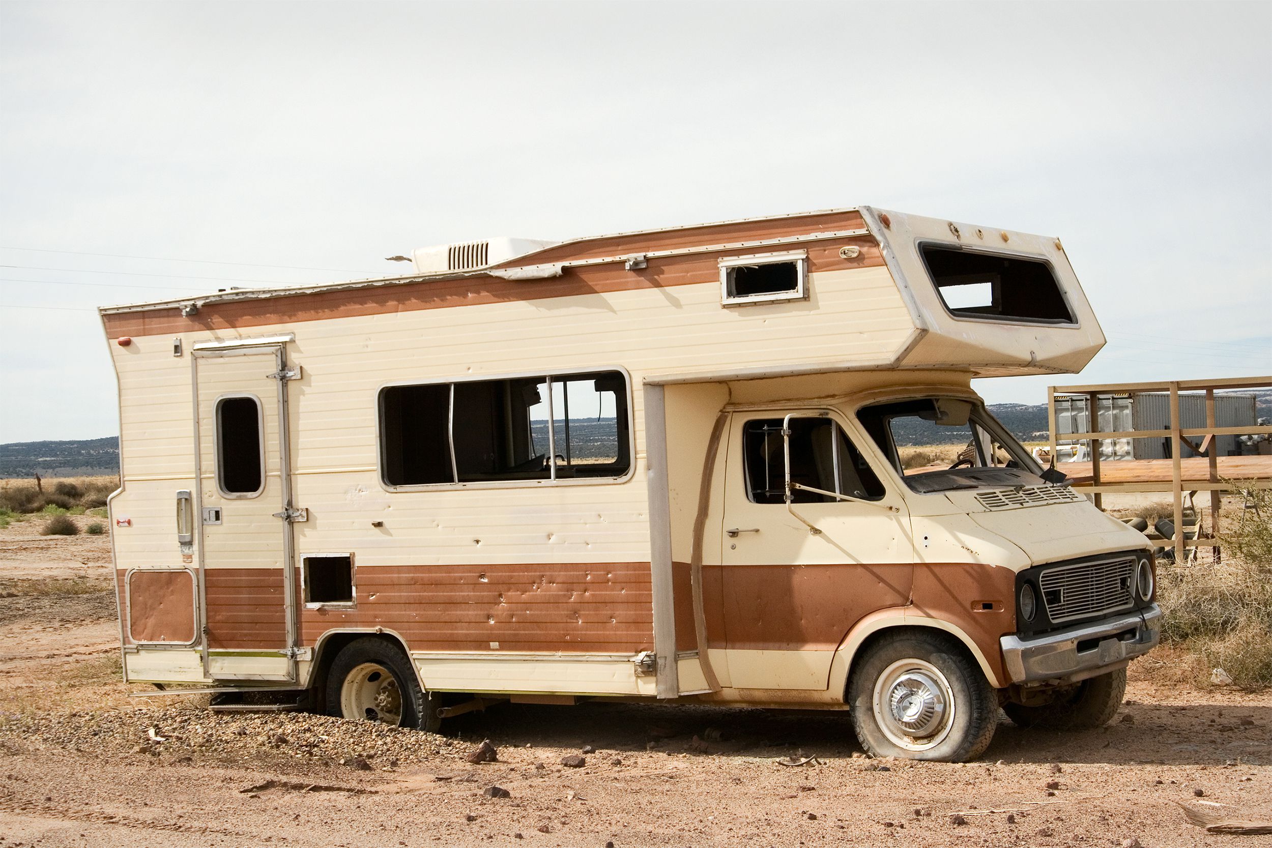 Side-view of an old abandoned beige and brown lined RV on dirt, dirt, junkyard, and light blue sky in the background