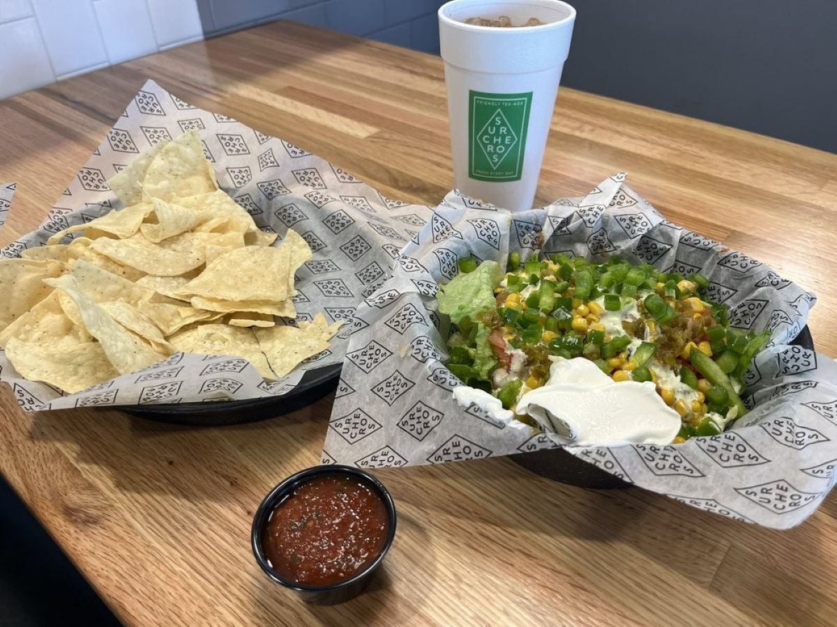 A basket of tortilla chips with salsa, a basket of loaded nachos topped with cheese, green onions, lettuce, and sour cream, and a white cup with a drink on a wooden table.