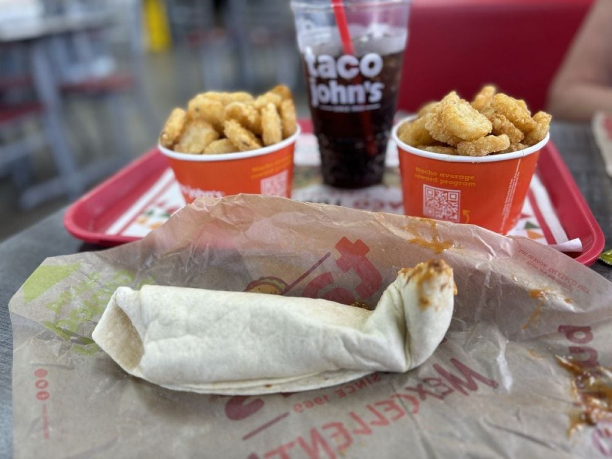 A wrapped burrito sits on paper next to two cups of seasoned potato bites and a fountain drink in a Taco John’s cup, all on red trays at a restaurant table.