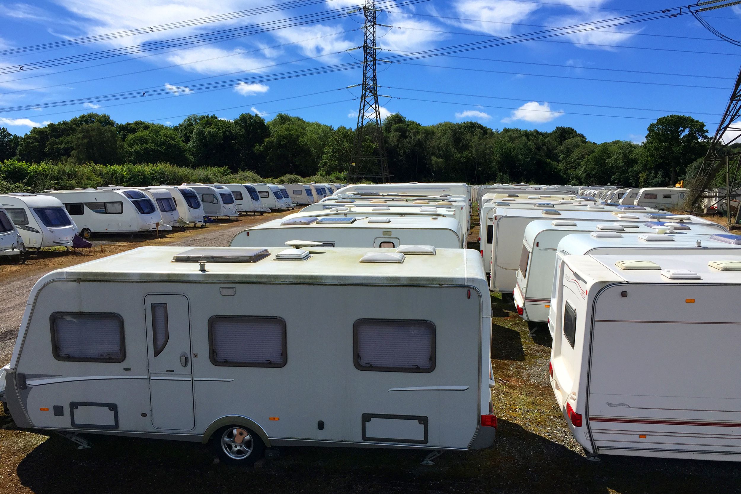 Three long rows of different white RVs stored in a lot at a dealership, trees, two transmission towers, and a blue sky on a sunny day