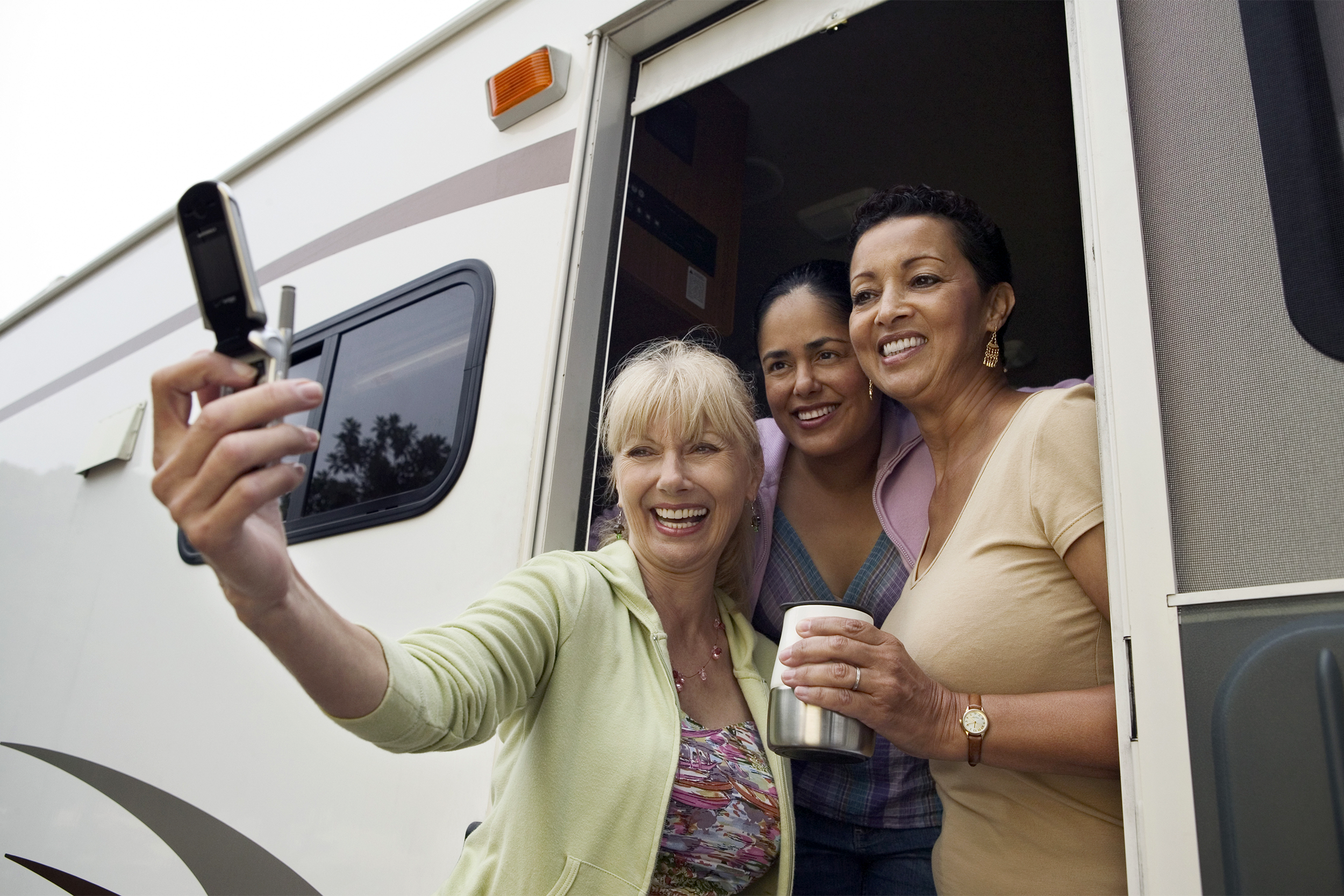 Three woman hanging out of the door of an RV, all smiling, taking a selfie, on a cell phone, on the right, side of white RV on the left, white background on the top left