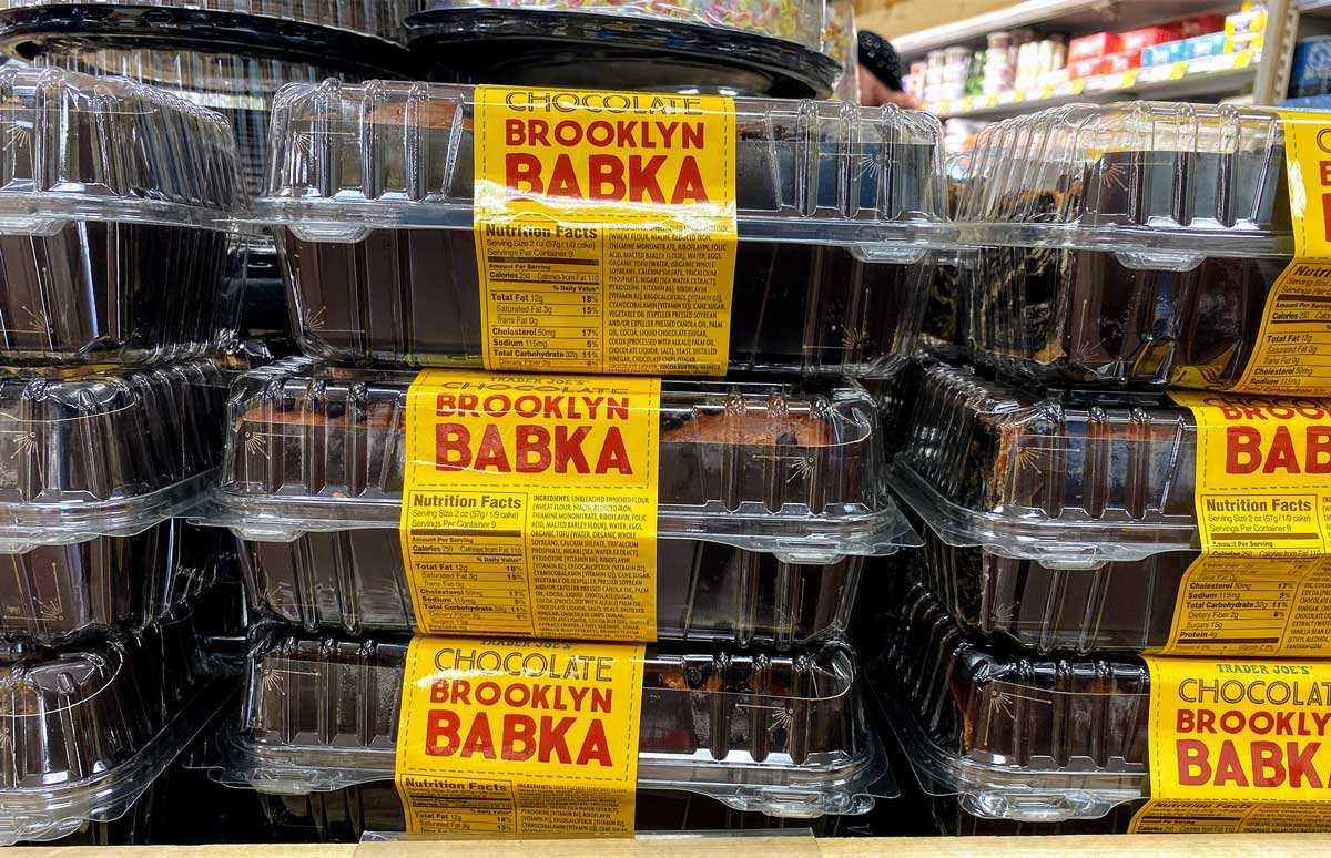 Plastic containers of chocolate Brooklyn babka, a favorite among Trader Joe's desserts, are stacked on a grocery store shelf. The yellow labels with red lettering stand out on each package, with more shelves and products visible in the background.