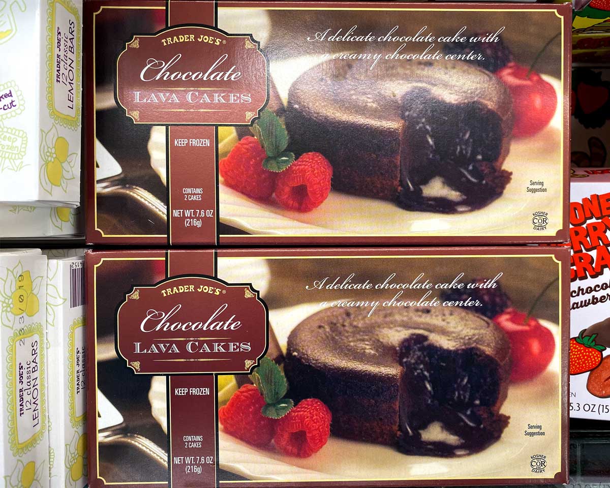 Boxes of Trader Joe's Chocolate Lava Cakes, part of the acclaimed Trader Joe's desserts lineup, show a molten-centered chocolate cake garnished with raspberries, a strawberry, and mint. The packaging highlights the dessert’s creamy chocolate center.