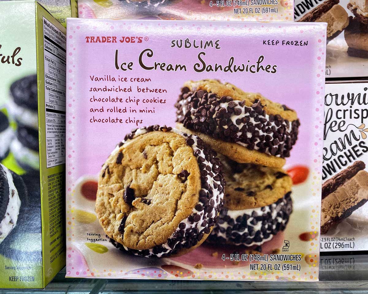 A box of Trader Joe’s Sublime Ice Cream Sandwiches, one of the standout Trader Joe's desserts, shows vanilla ice cream between chocolate chip cookies rolled in mini chocolate chips. The box contains four sandwiches against a pink background.