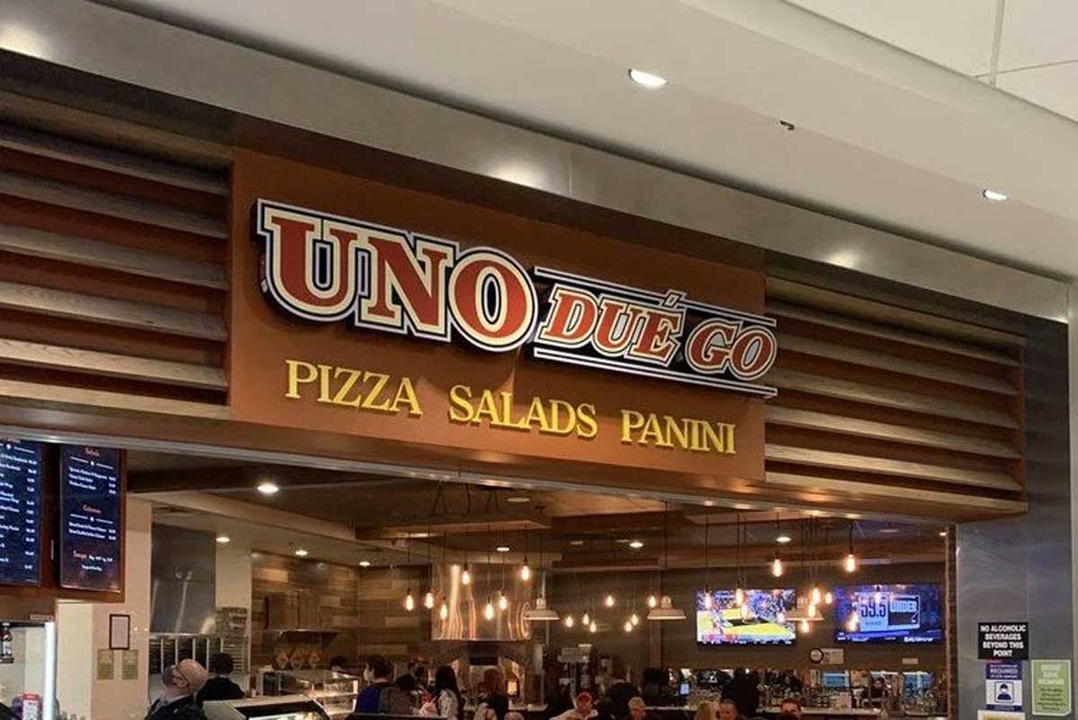 Restaurant storefront with a sign reading "UNO DUE GO" and "PIZZA SALADS PANINI" underneath. The interior shows a counter, hanging lights, menu boards, and customers ordering food—unlike some failed pizza chains, this spot is bustling.