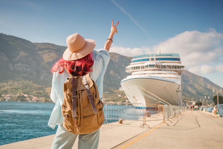 A woman with a backpack and hat stands on a dock, facing a large cruise ship. She raises her hand in a peace sign, with mountains and blue sky in the background.