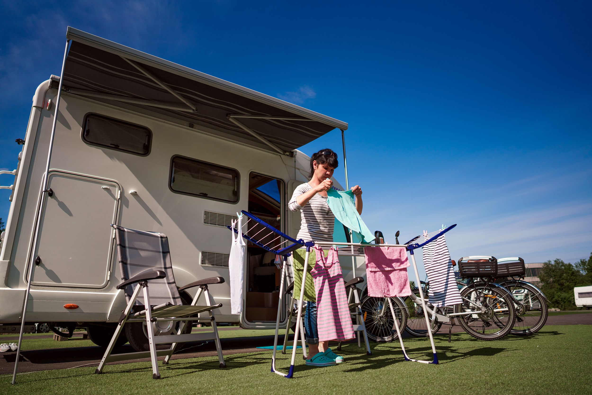 Woman hanging clothes on hanger on lawn in front of an RV, two outdoor folding chairs surround her, two bikes on the right, a blue sky in the background