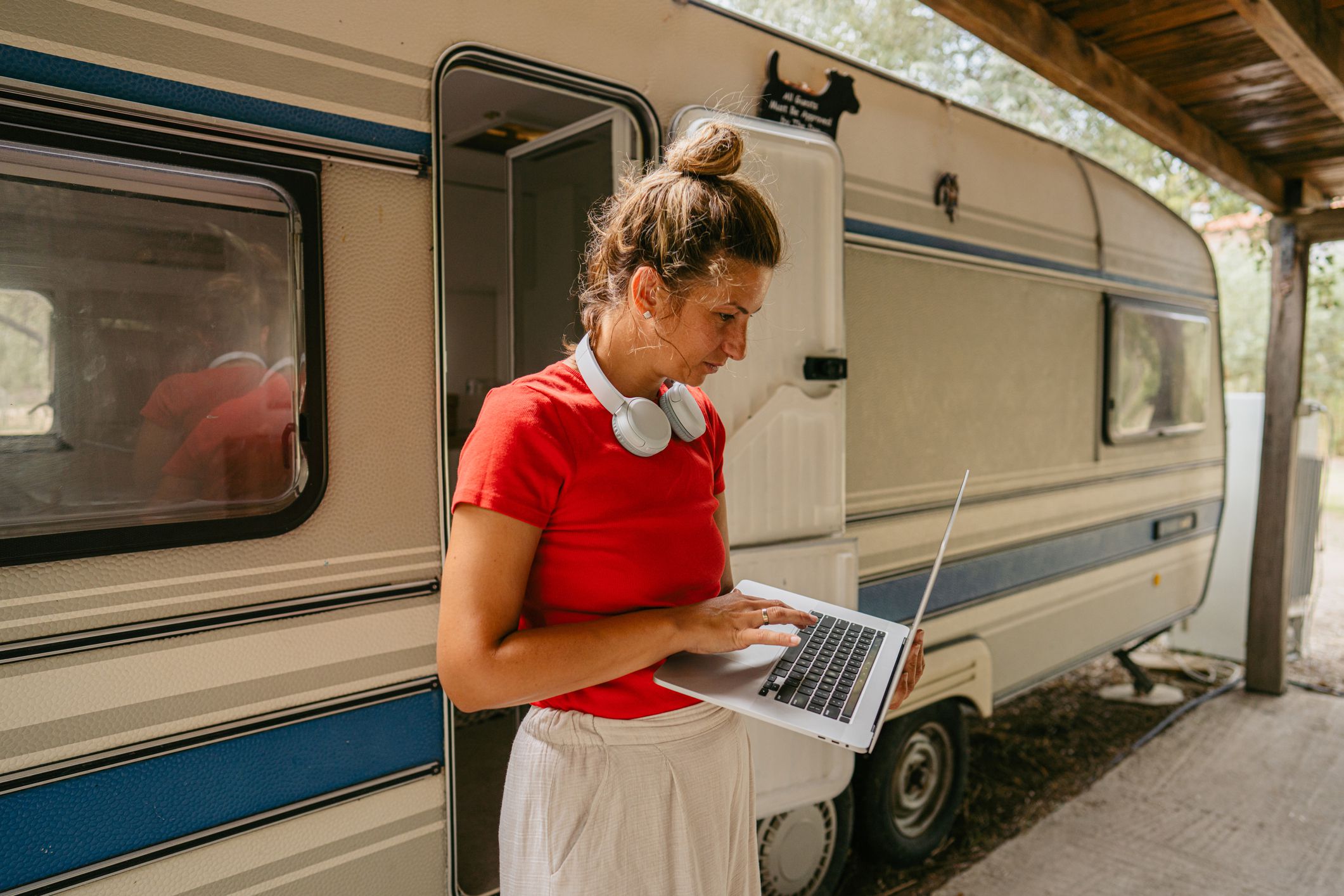 Woman holding a MacBook while standing, on a porch in front of an RV, trees and lawn in the background on the top and right