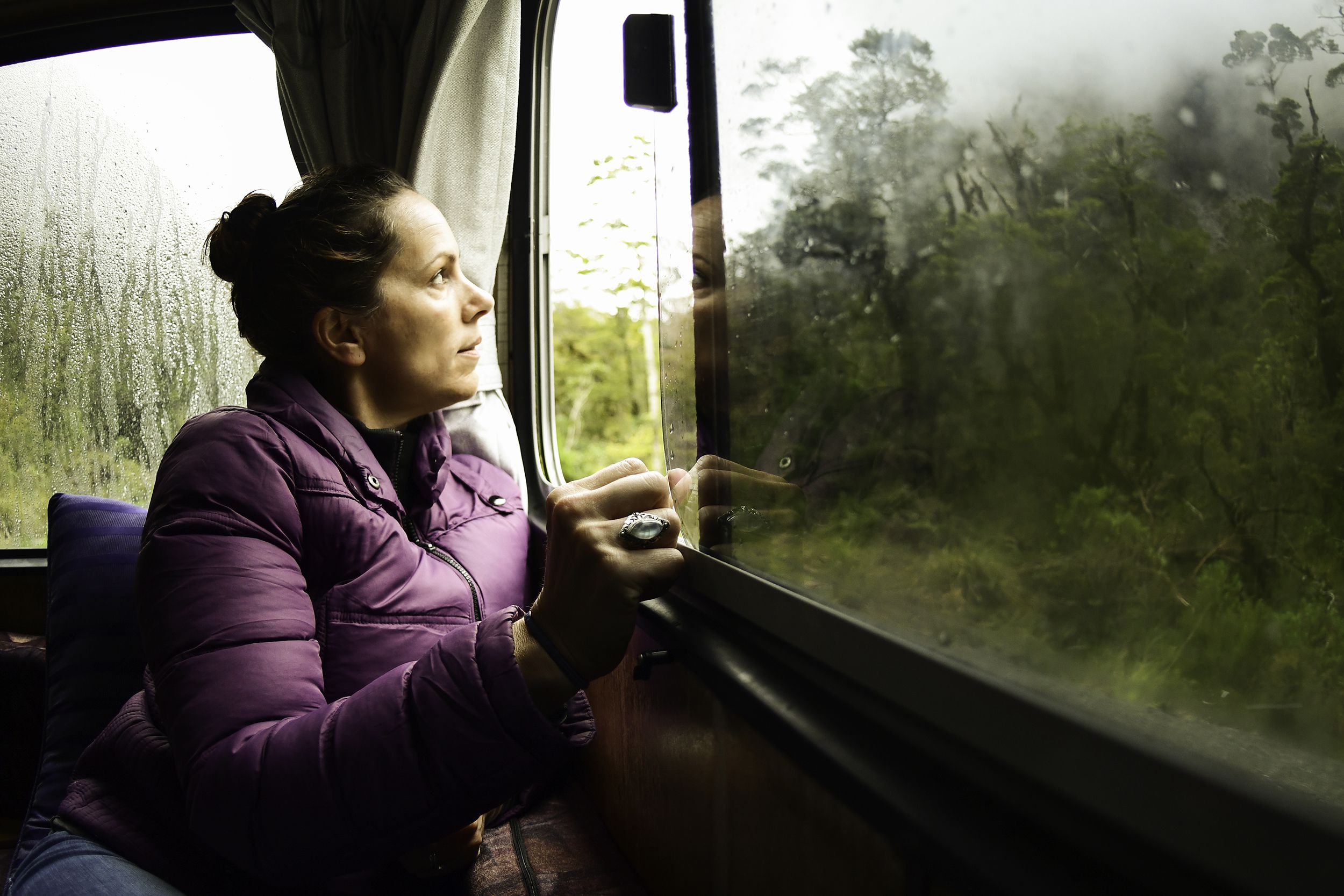 Woman sitting inside an RV, opening the window, looking outside at trees and forest during a rainstorm, shadow, she is wearing a purple puffer jacket
