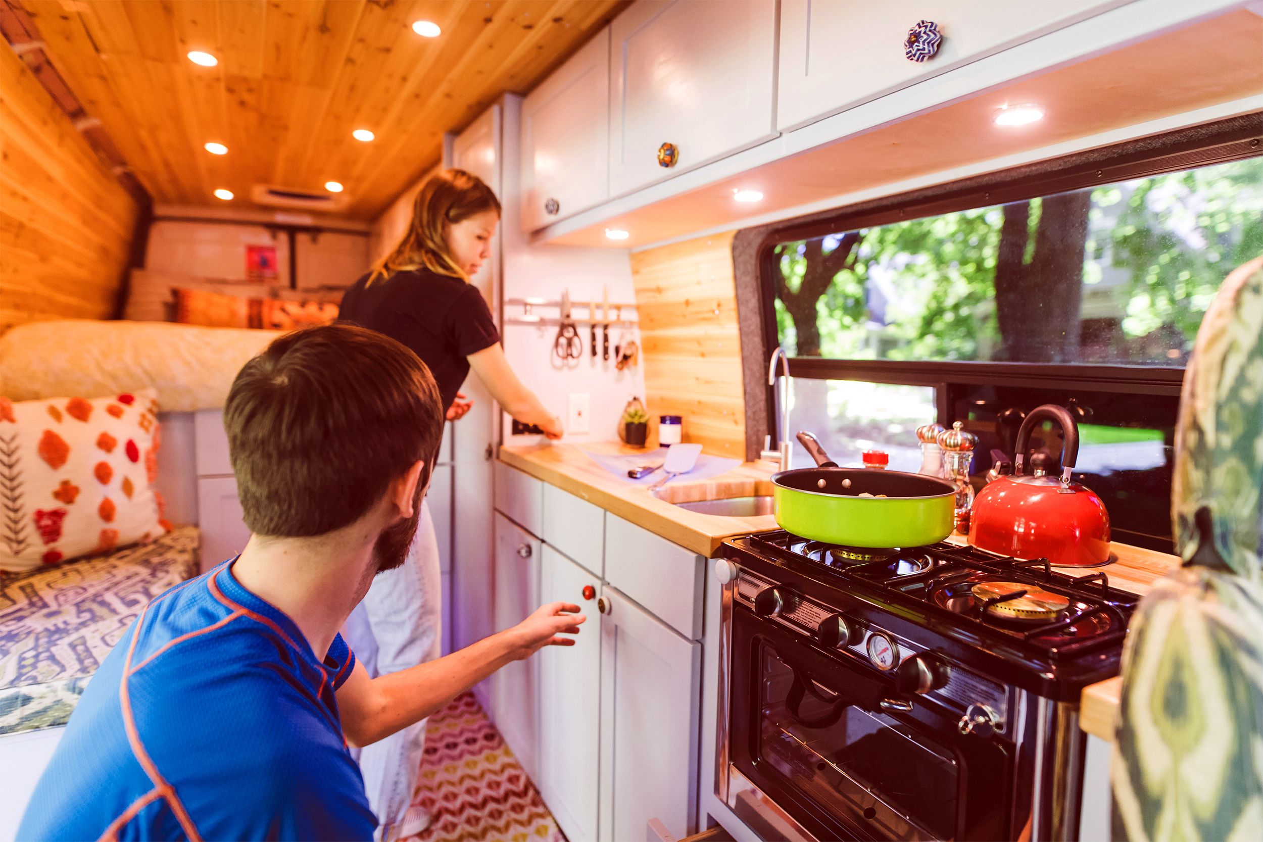 Young couple in the kitchen of a modern RV, cooking and preparing food, trees and greenery on a sunny day through the kitchen window