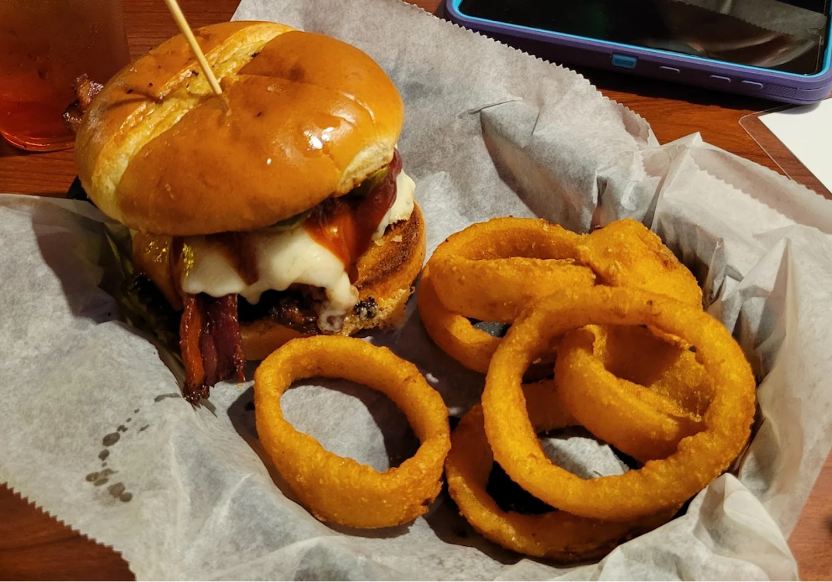 A cheeseburger with bacon, lettuce, and tomato sits on wax paper in a basket next to several golden-brown onion rings. A drink and a tablet are partially visible in the background.