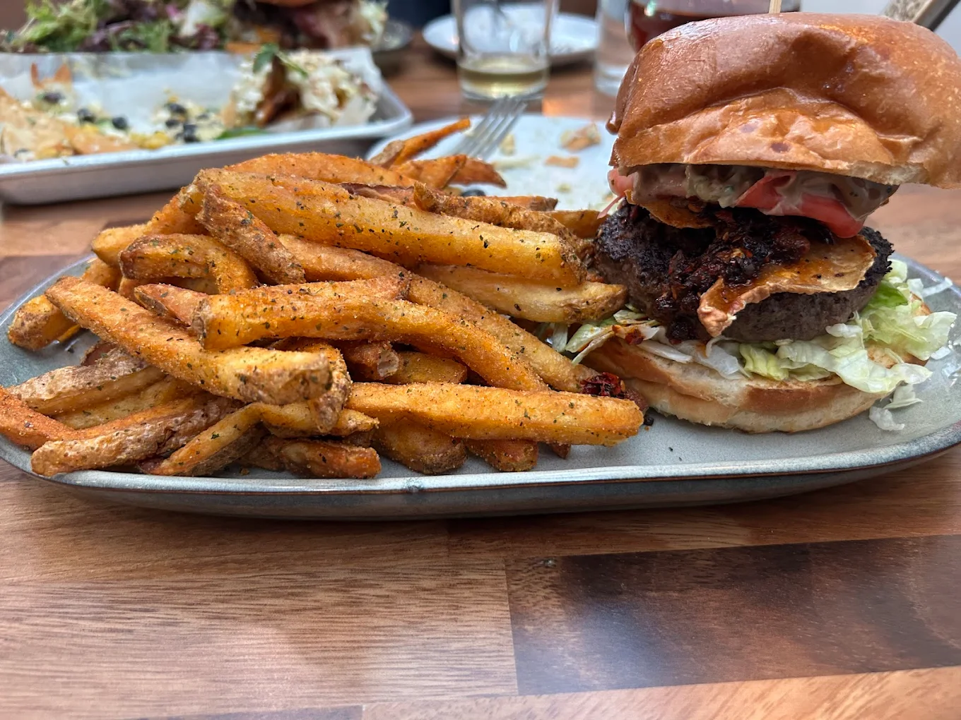 A metal tray with seasoned French fries and a burger with lettuce, tomato, and pickles on a brioche bun, on a wooden table. Other plates and glasses are blurred in the background.