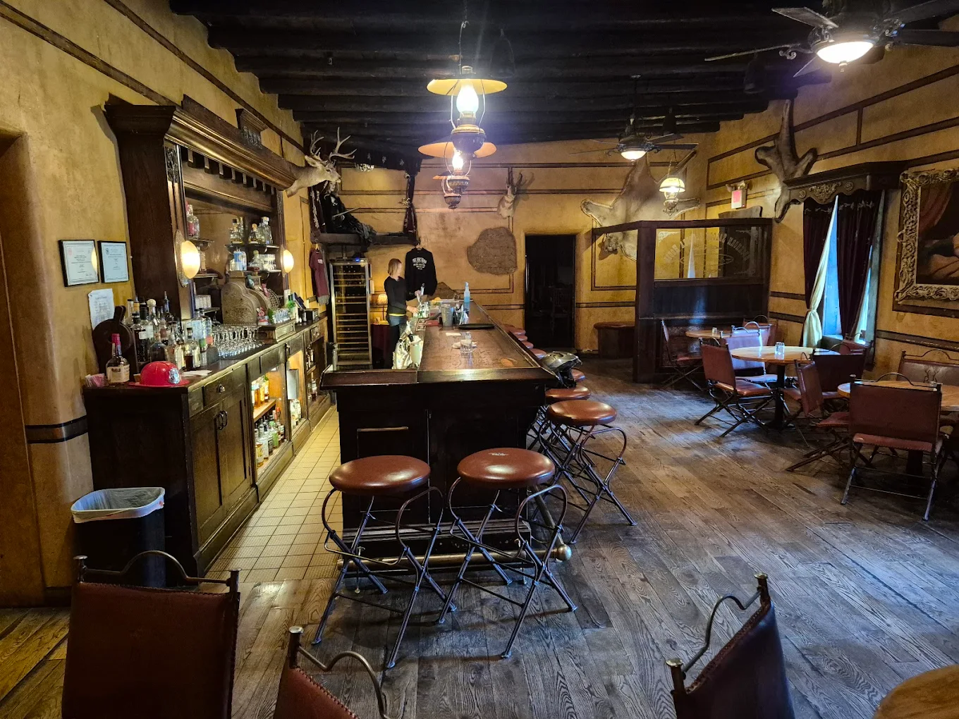 A rustic bar with wooden floors and vintage decor, featuring a long wooden counter with stools, shelves of bottles, and several tables and chairs. Antlers and old photos decorate the walls, creating a cozy, old-fashioned atmosphere.