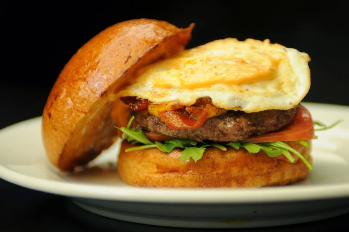 A close-up of a gourmet burger with a fried egg on top, lettuce, tomato, and a beef patty inside a toasted bun, served on a white plate against a black background.