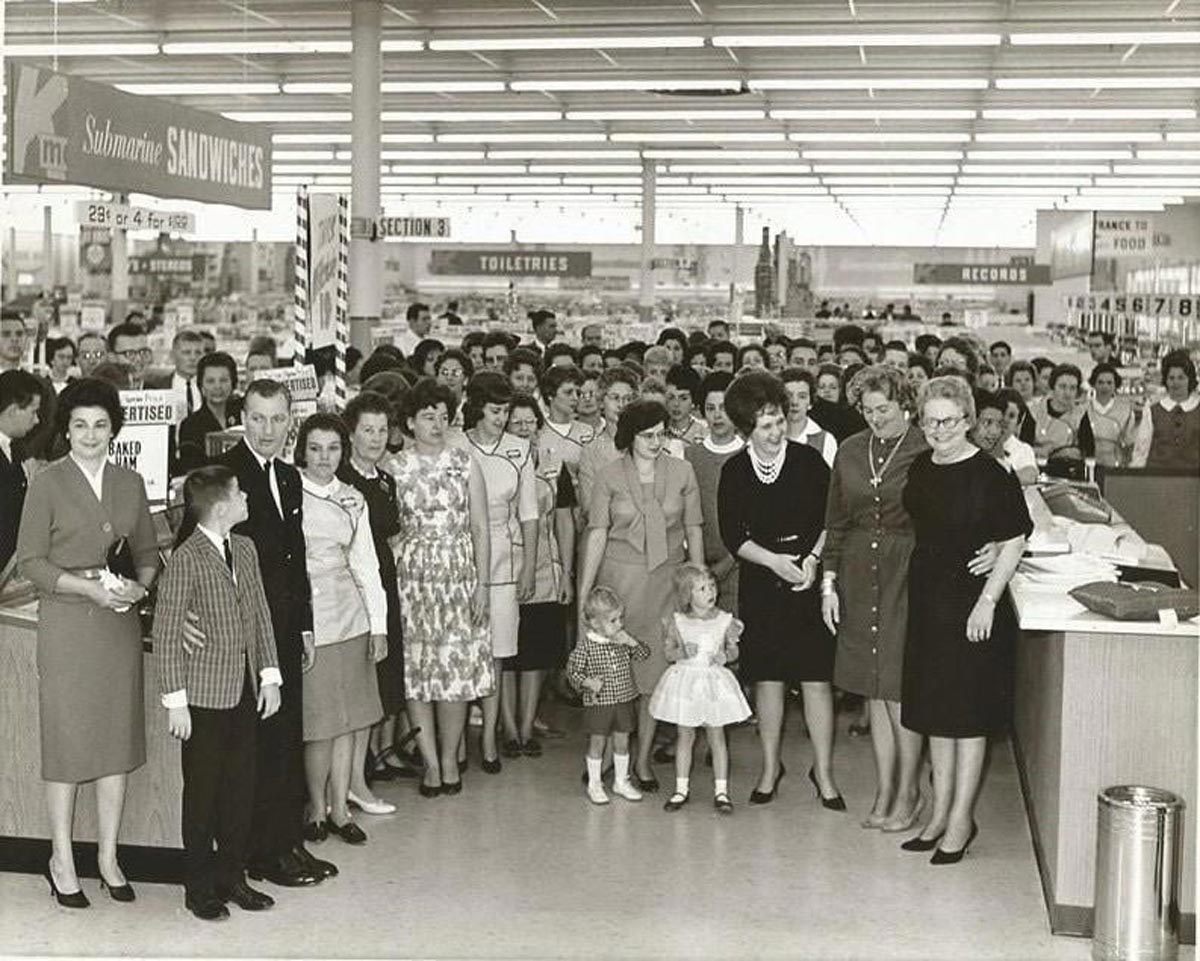 A large group of people, mostly women and children, stand together inside a brightly lit department store, posing for a photo. Store signs for sandwiches, records, and toiletries are visible in the background.