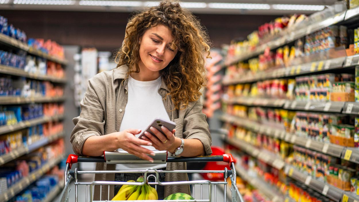 A woman with curly hair pushes a shopping cart with bananas and watermelon in a grocery store aisle while looking at her smartphone and smiling. Shelves stocked with products line both sides of the aisle.