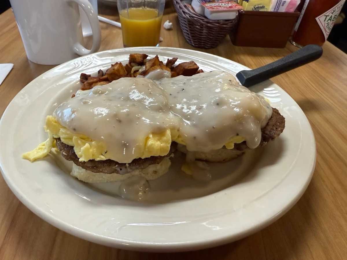 A plate of breakfast food featuring two biscuits topped with sausage patties, scrambled eggs, and white gravy, served with a side of diced, fried potatoes. A mug, orange juice, and condiments are visible in the background.