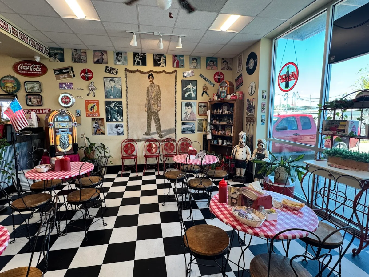 A retro diner with black-and-white checkered floor, red chairs, round tables with red checkered tablecloths, Elvis Presley mural, vintage memorabilia on the walls, and large windows bringing in sunlight.