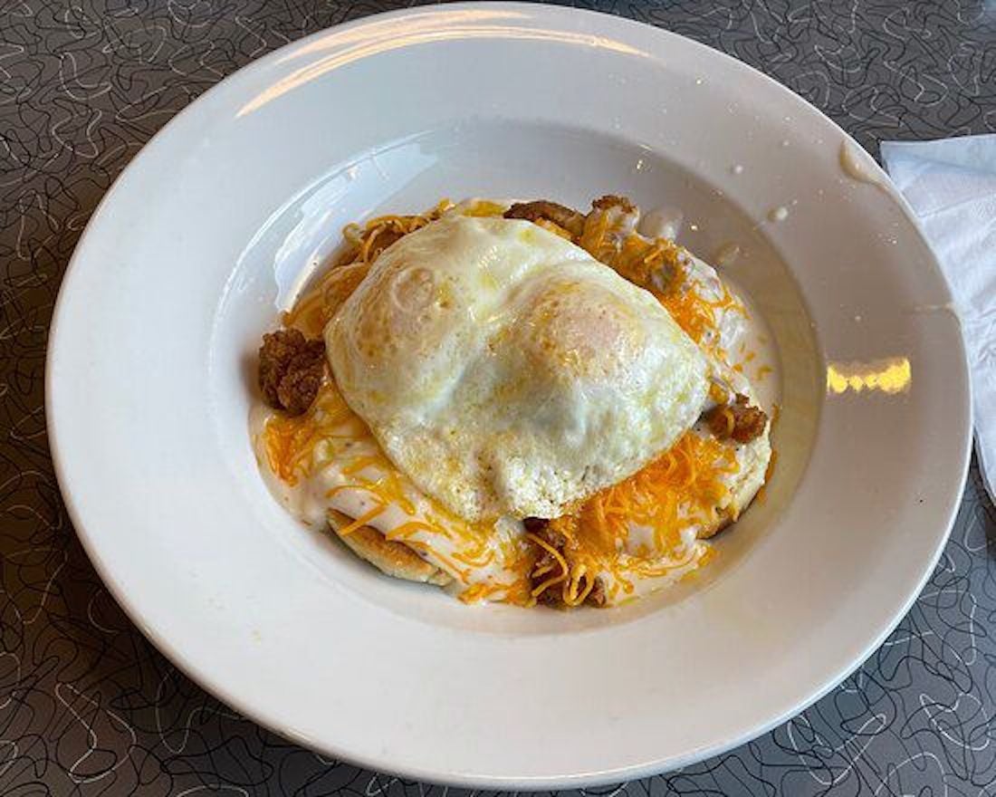 A white plate holds two sunny-side-up eggs atop a biscuit covered in white gravy, shredded cheese, and pieces of fried chicken, with a napkin on the side.