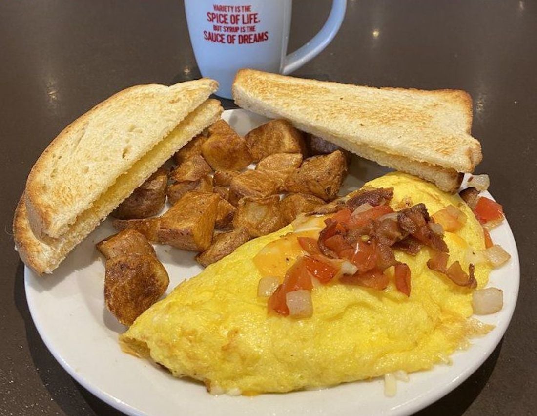 A breakfast plate with an omelet topped with diced tomatoes, onions, and bacon, served with golden brown home fries and two slices of toasted bread. A coffee mug is in the background.