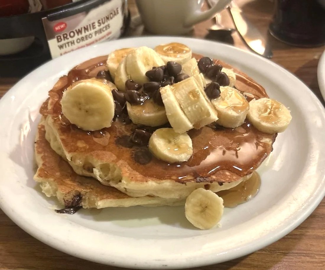 Two Denny's pancakes topped with banana slices, chocolate chips, and syrup are served on a white plate. A menu featuring a brownie sundae is visible in the background.