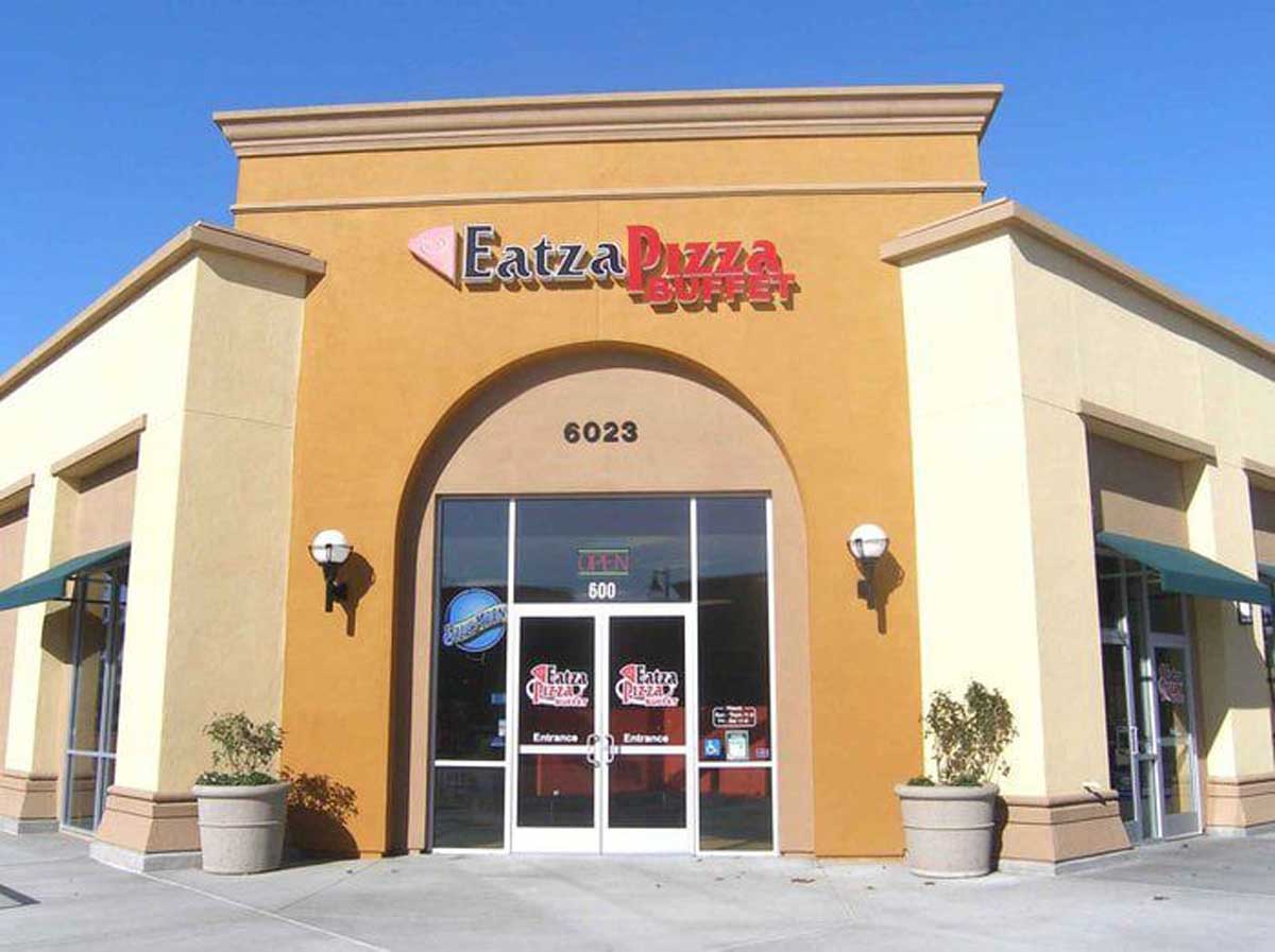A tan and yellow restaurant building with an arched entrance labeled “Eatzza Pizza Buffet.” Once one of the failed pizza chains, its glass doors display the restaurant’s logo, with large potted plants flanking the entrance.