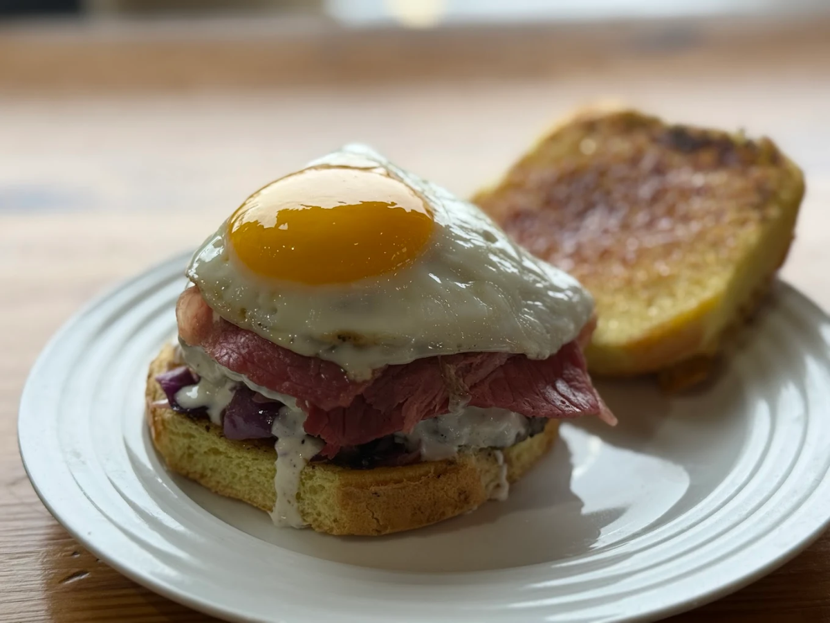 An open-faced sandwich on a white plate, topped with sliced meat, creamy sauce, purple cabbage, and a sunny-side-up egg. The other half of the toasted bun is placed to the side.