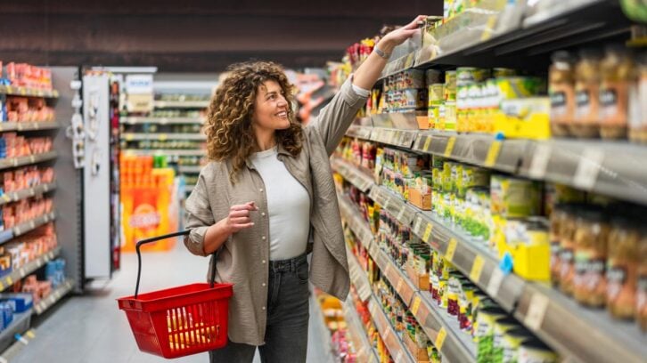 A woman with curly hair smiles as she reaches for an item on a high supermarket shelf, holding a red shopping basket in her other hand. She stands in an aisle lined with various packaged goods.