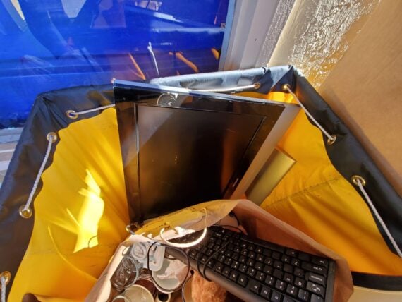 A computer monitor, keyboard, and various small items are discarded in a yellow bin, next to a window with sunlight shining in.